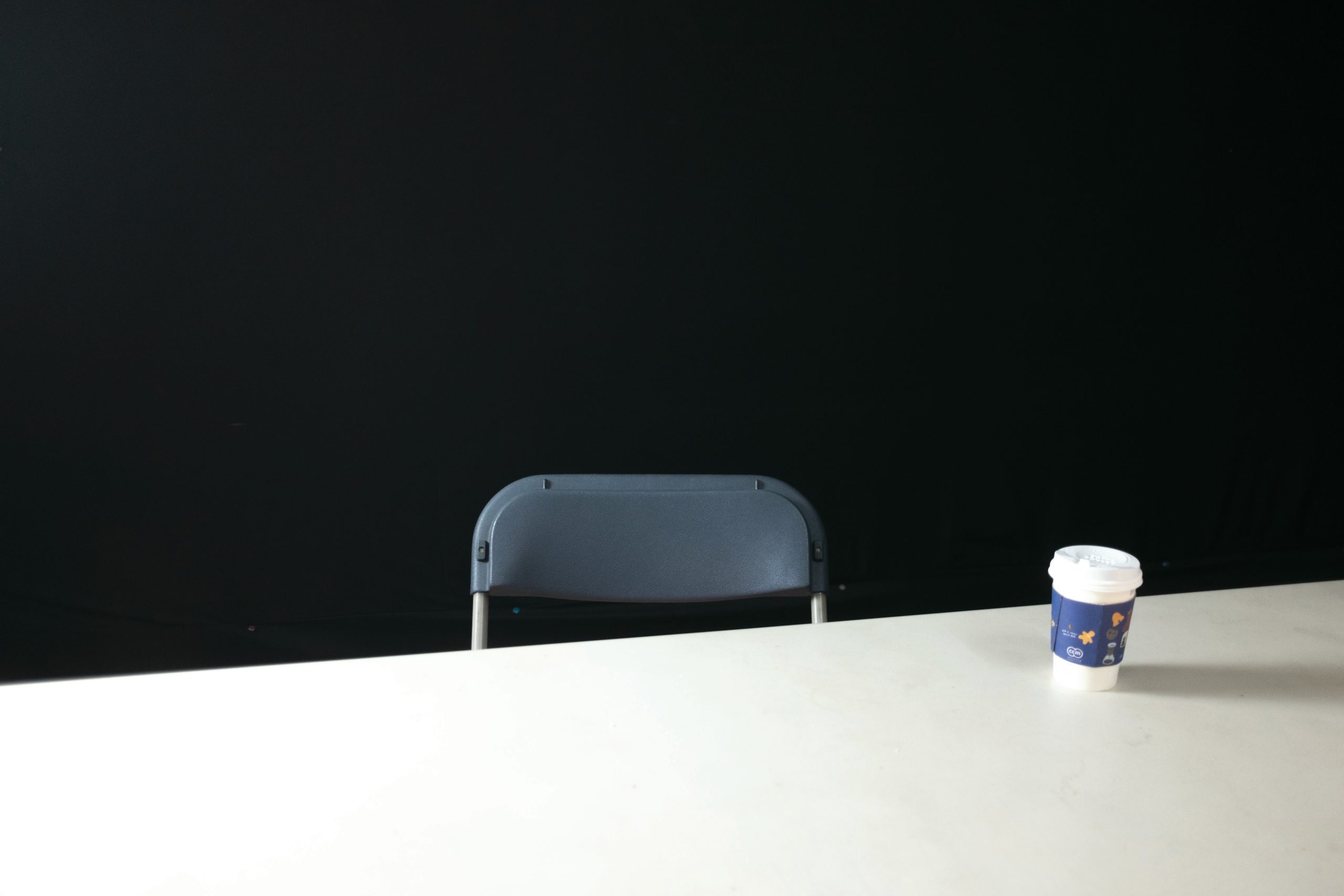 Mesa con un café para llevar y una silla plegable detrás de la mesa, con un fondo oscuro que recuerda a una sala de interrogatorios